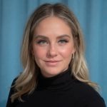 Portrait of a woman with wavy blonde hair, wearing a black top and gold earrings, set against a blue background. She has a calm and confident expression.