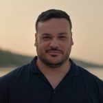 A man with short dark hair and facial hair stands outdoors at sunset, wearing a dark shirt. The background shows a soft-focus coastline and sky.